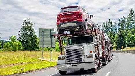 White semi car hauler carrying a red SUV on an open trailer along a highway with trees