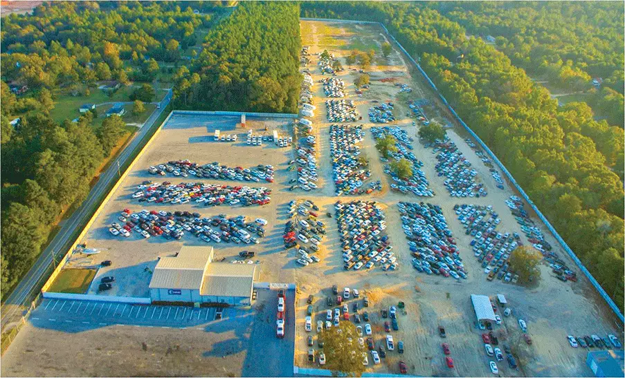 Aerial view of a large auto auction yard with hundreds of vehicles parked in organized rows surrounded by trees
