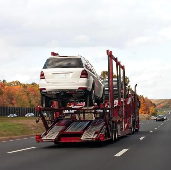 Red open auto carrier transporting multiple vehicles on a highway with autumn scenery in the background