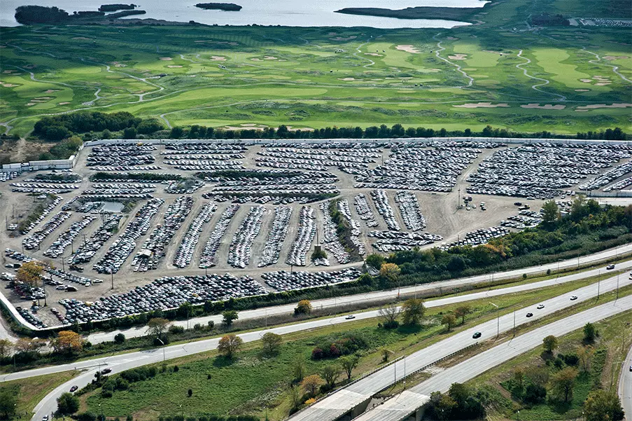 Aerial view of a large auto auction yard filled with rows of vehicles near a highway and green landscape in the background