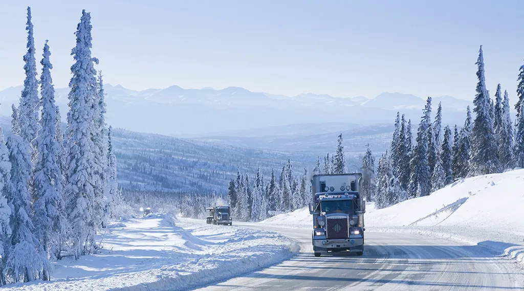 Semi truck driving on a snow covered highway through a winter mountain landscape with frost covered trees