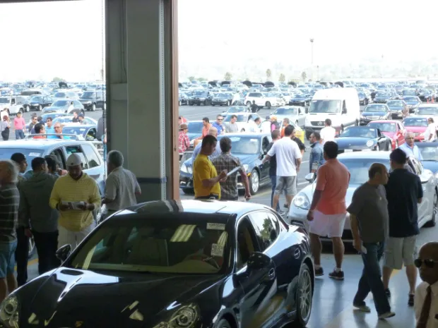 Car auction scene with buyers inspecting vehicles inside a large warehouse and a crowded parking lot outside