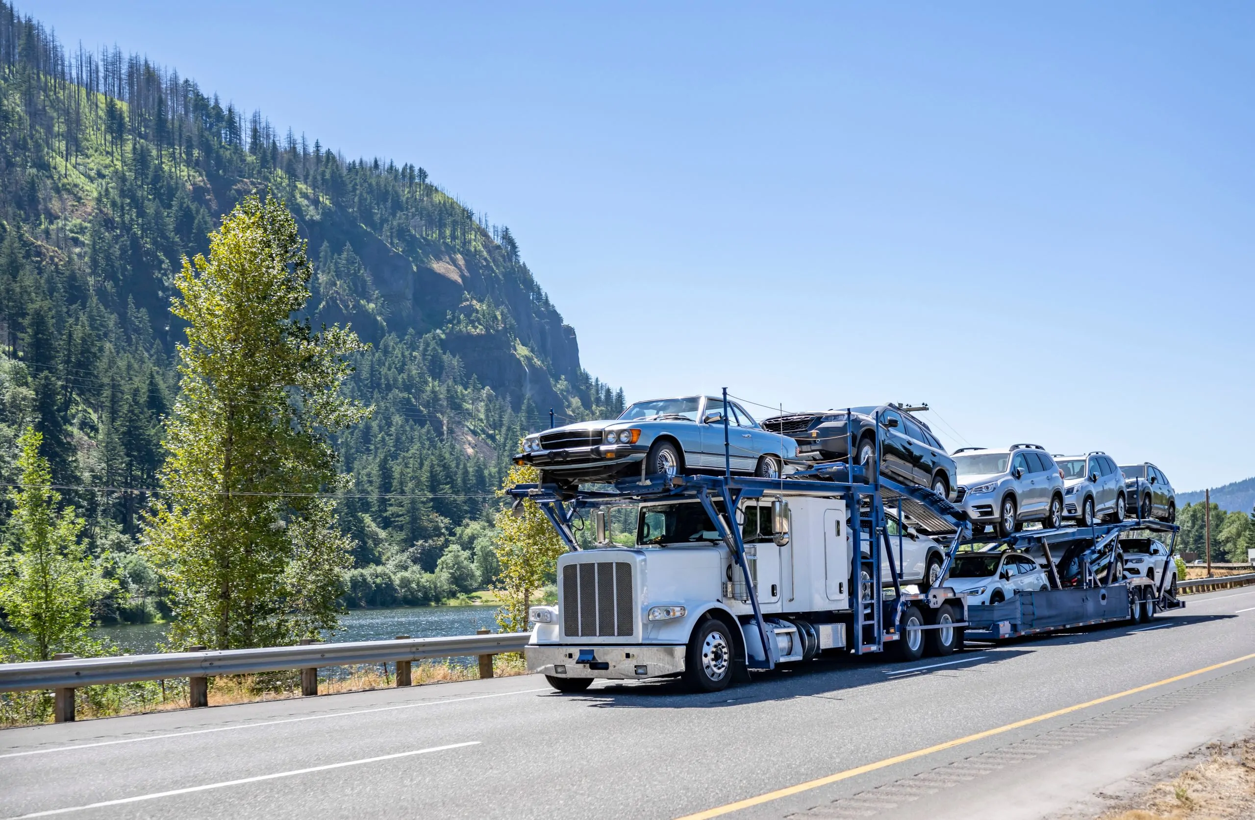 Open auto transport truck hauling multiple vehicles on a highway beside mountains, trees, and a river in a scenic landscape