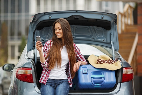 college student sitting on car trunk with luggage checking phone before move